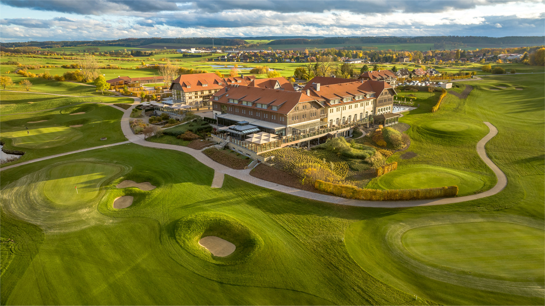 Golf course with a clubhouse surrounded by greenery and a cloudy sky.